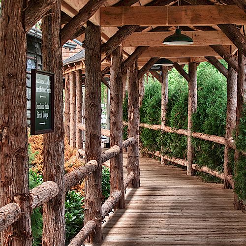 A rustic wooden walkway covered by a wooden roof, surrounded by greenery. The path is enclosed with wooden railings, leading through a scenic area.