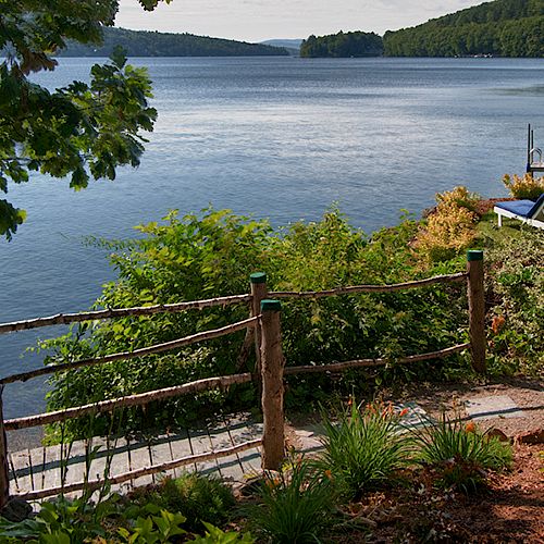 The image shows a serene lakeside view with a rustic fence, lush greenery, lounge chairs by the water, and a backdrop of trees and hills.