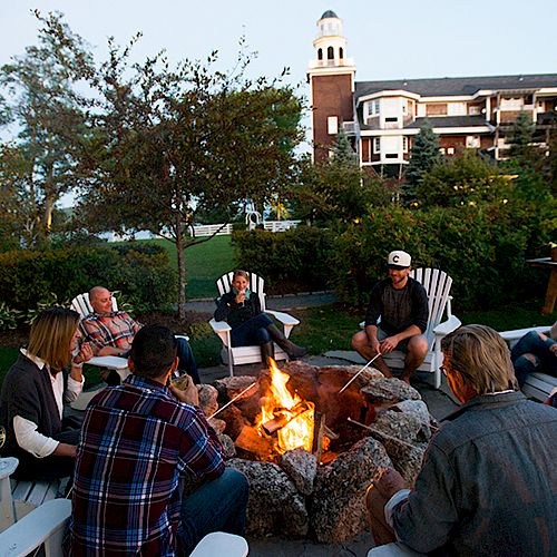 A group of people is gathered around a fire pit in a garden. They are sitting on white chairs and appear to be roasting marshmallows.