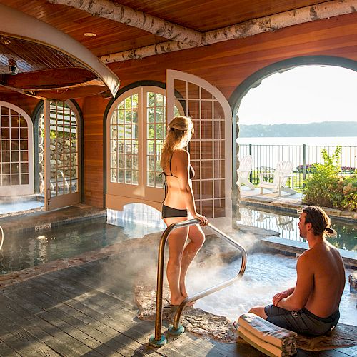 A man and woman enjoying a hot tub in a wooden indoor spa area with open doors providing a scenic view of a lake and mountains outside.