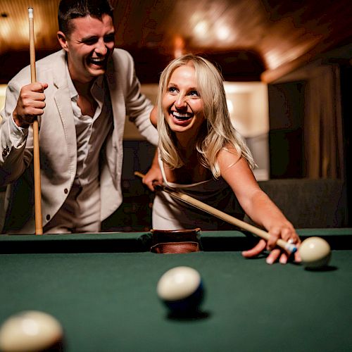 Two people are joyfully playing pool, one aiming while the other holds a cue stick, in a warmly lit room.