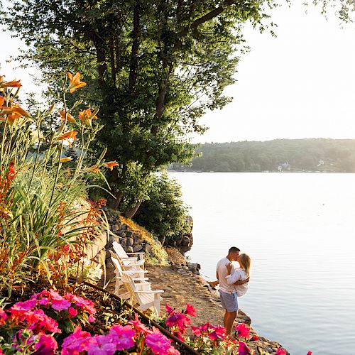 A couple embraces by a lake with colorful flowers in the foreground, trees around, and a building nearby under a serene sunset sky.