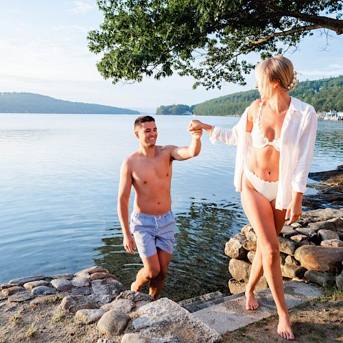 A couple is enjoying a sunny day at a lake, with the man helping the woman out of the water onto a stone path.