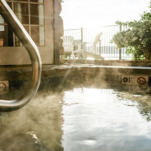 The image shows a steaming hot tub with handrails, warning signs, surrounded by a lush garden and chairs in the background.