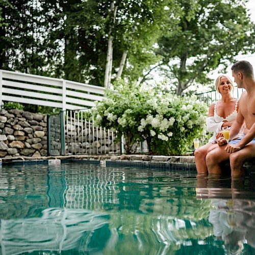 A man and a woman sit at the edge of a pool, chatting and enjoying the outdoors, surrounded by greenery and stone walls.