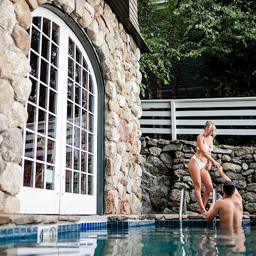 A woman in a swimsuit helps a man out of a pool with stone walls and large windows in the background. Trees and white fence are visible.