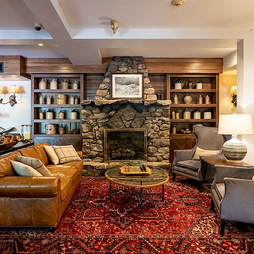 A cozy living room with a stone fireplace, leather sofa, armchairs, and a red patterned rug. Shelves with decor items flank the fireplace.