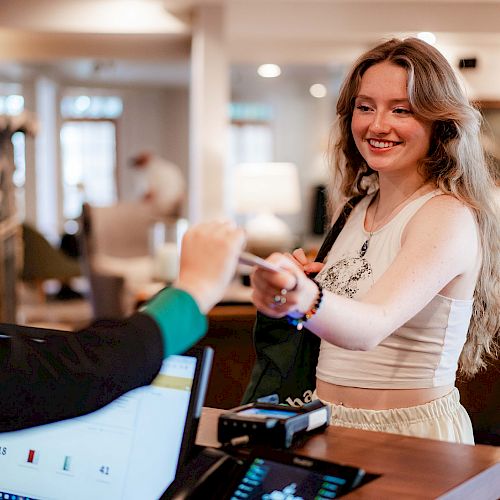 A smiling woman is interacting at a reception desk, possibly checking in or making a payment, with a person behind the counter handing her a key.