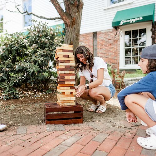 Three kids are playing a giant outdoor Jenga game in a garden setting near a brick building and greenery, enjoying the warm weather.