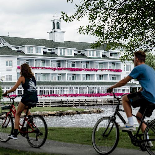 Two people are riding bicycles on a path beside a body of water, with a large white building with floral balconies in the background.