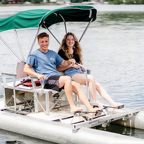 A couple is sitting and smiling on a small pedal boat with a green canopy, floating on a calm body of water.