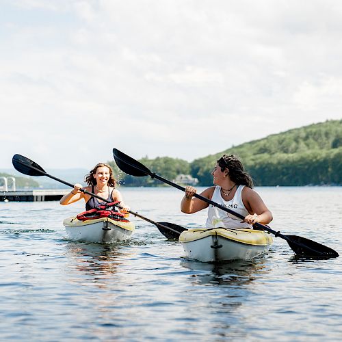 Two people are kayaking on a calm lake with trees and hills in the background, enjoying a sunny day outdoors.