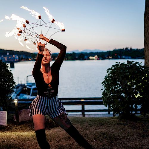 A person performs a fire dance with a lake and boats in the background, during twilight. They are holding multiple flaming props, engaging in an energetic dance.