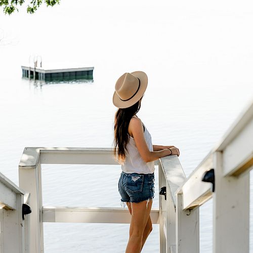 A person wearing a hat and shorts stands on a staircase by a calm body of water, looking towards a floating platform.