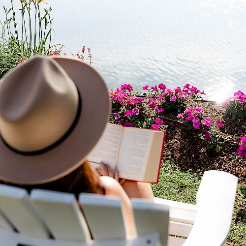 A person wearing a hat sits in a white chair by a body of water, reading a book, with flowers in the background.