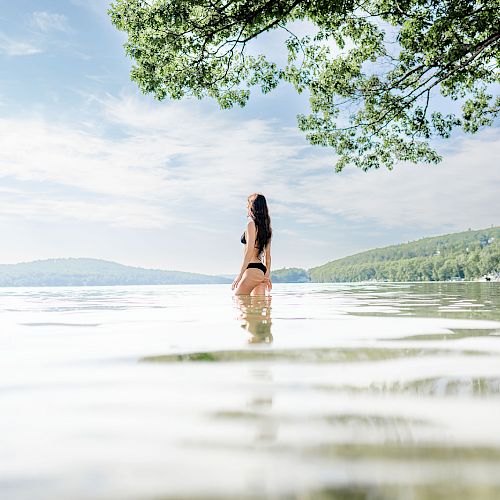 A person stands in shallow water at a scenic lake, with lush green hills and a partially cloudy sky in the background, under a tree branch.