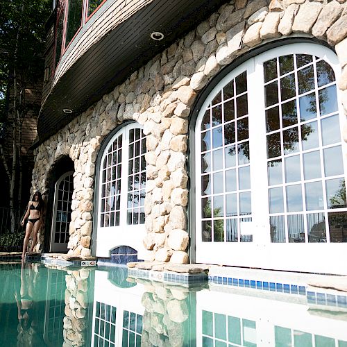 A stone building with large arched windows is reflected in a swimming pool. A person is standing near the pool and the surrounding greenery.