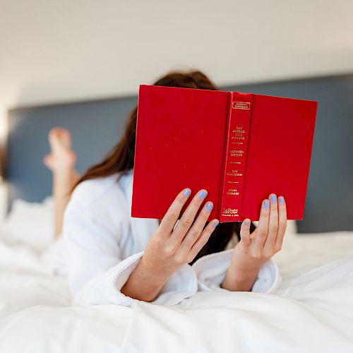 A person is lying on a bed, reading a red book. They are holding the book with both hands, and their face is hidden behind it, ending the sentence.