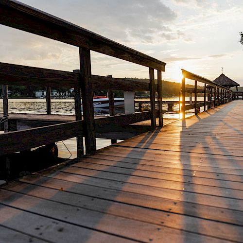 The image shows a wooden boardwalk by a lake, with the sun setting, casting long shadows. There are boats and a gazebo in the background.