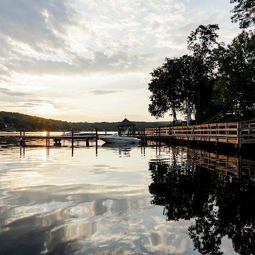 A peaceful lakeside scene at sunset with a dock, boats, trees, and calm waters reflecting the sky and surroundings.