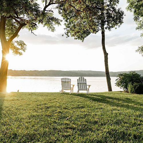 This image features two white Adirondack chairs on a grassy shore overlooking a serene lake, framed by trees, with the sun setting in the background.