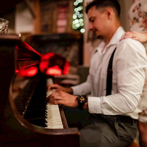 A man is playing a piano while a woman stands beside him, resting her hand on his shoulder. There is a drink on the piano.