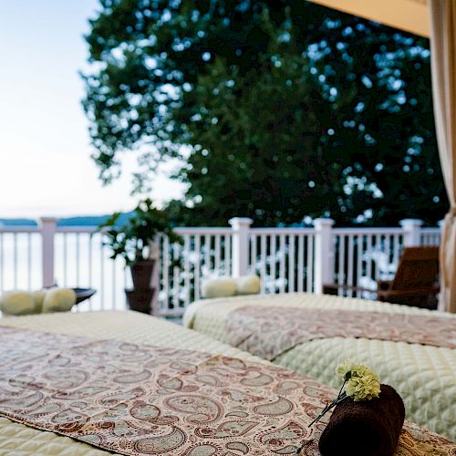 The image shows two massage tables on a balcony overlooking a serene, tree-lined waterfront, with plants and rolled towels enhancing the tranquil setting.