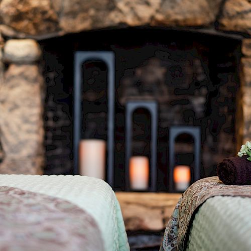 The image shows a cozy stone fireplace with candles and two beds covered with blankets, one topped with a rolled towel and a flower.