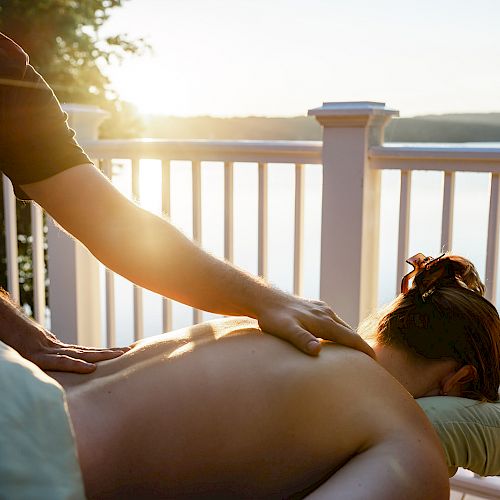 A person is receiving a back massage on an outdoor deck overlooking a serene lake at sunset, with a massage therapist working on their back.