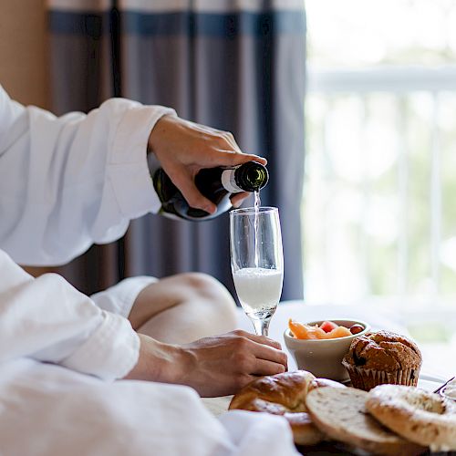 A person in a robe is pouring a drink into a glass with breakfast items including bread, fruit, and a muffin on a tray in front of them.