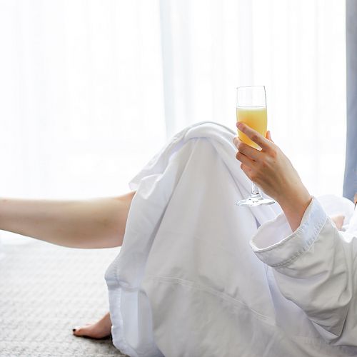 A person in a white robe is relaxing, holding a drink, likely a mimosa, with legs stretched out in a cozy room with light curtains.