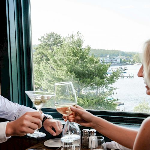 Two people clinking glasses at a table near a window with a scenic view of trees and water in the background.