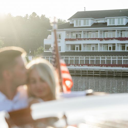 A couple enjoying a moment on a boat with a large building and American flag in the background on a sunny day.