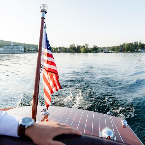 A person's hand rests on a boat with an American flag at the stern, sailing on a lake surrounded by trees and distant hills.