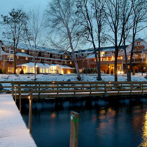 An illuminated building next to a snowy dock and water, surrounded by trees in a winter setting, under a twilight sky.