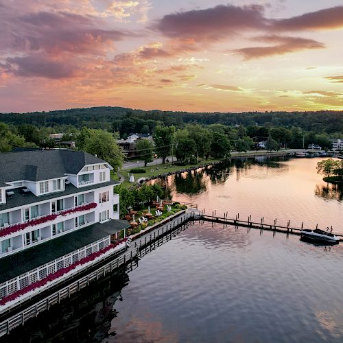 A lakeside building with a docked boat, surrounded by water and greenery, under a colorful sunset sky with scattered clouds.