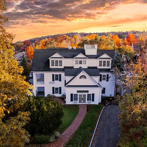 A charming white house sits among trees with autumn foliage, under a colorful sky at sunset, with a curved path leading to the front door.