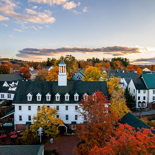 An aerial view of a picturesque town with white buildings, a central clock tower, and surrounding autumn-colored trees under a partly cloudy sky.