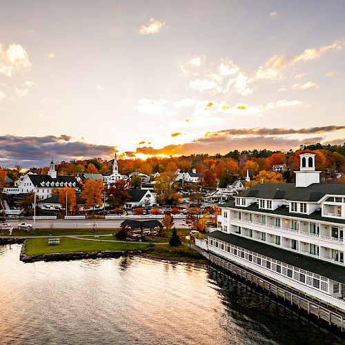 A serene lakeside town during sunset with autumn foliage, traditional buildings, and a white multi-story building by the water’s edge.