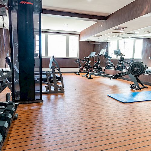 The image shows a gym with dumbbells, rowing machines, stationary bikes, and a blue exercise mat on a wooden floor, with mirrors on the walls.