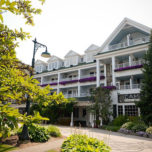 The image depicts a charming multi-story building, surrounded by greenery and flowers, with balconies adorned with purple flowers.