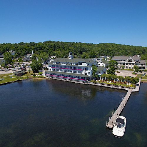 A waterfront building with a dock, a boat, and trees in the background under a clear blue sky, seen from above the water.