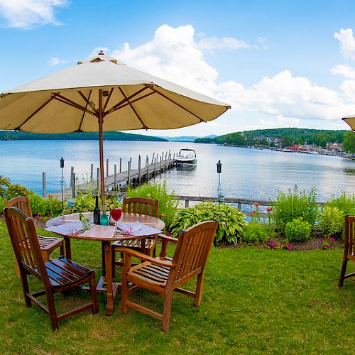 Outdoor seating area with wooden tables and chairs under large umbrellas, overlooking a scenic lake with a dock and boat in the background.