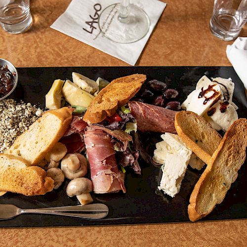 A charcuterie board with assorted meats, cheeses, bread, mushrooms, and jam, served on a black slate with rolled napkins and drinks on the side.