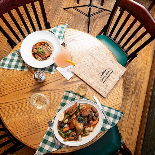 A round wooden table with two plates of food, drinks, and a menu on a wooden floor, surrounded by three chairs and green checkered napkins.