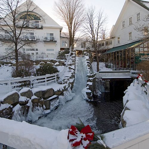 A snowy outdoor scene shows a frozen waterfall with adjacent white buildings, decorated with festive greenery and red bows, under a clear sky.