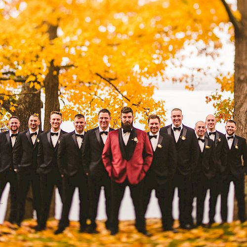 A group of men dressed in formal attire, with one in a red suit, pose outdoors among autumn trees with golden leaves, in a picturesque setting.