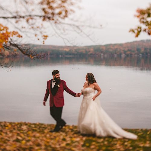 A couple in wedding attire walks hand in hand by a serene lake with autumn foliage in the background, showcasing the beauty of the season.