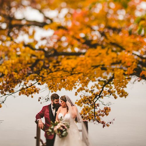 A couple in wedding attire poses under a tree with vibrant fall foliage by a lake. The scene is romantic and picturesque, showcasing autumn beauty.