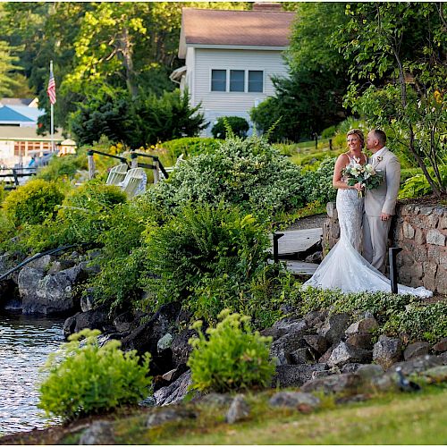 A bride and groom stand near a stone wall by a waterfront surrounded by greenery and houses, posing for a photo on their wedding day.